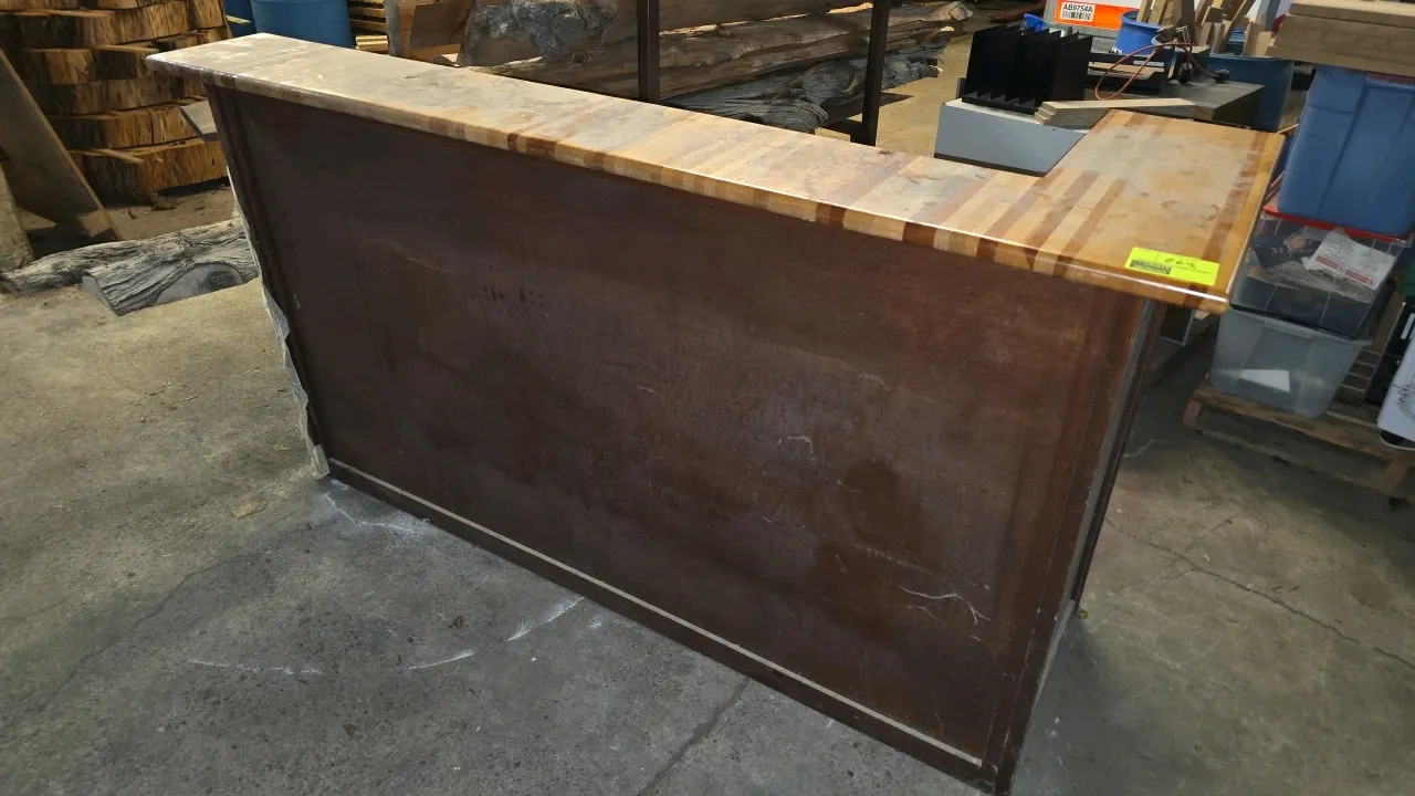 Wood L-shaped bar counter. Features butcher-block style top surface, dark wood finish front paneling, and an open interior frame. 86"L x 46"W.