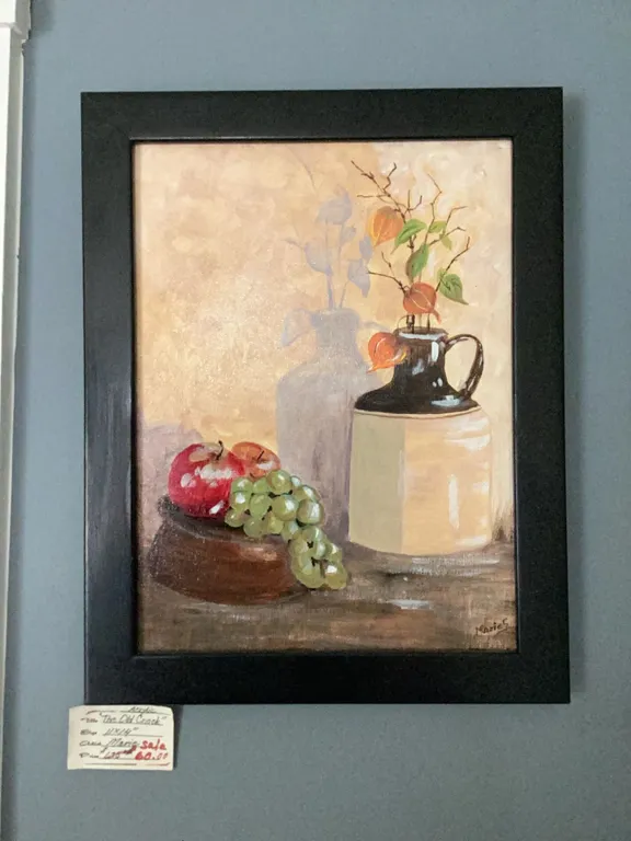 Framed still life painting featuring a cluster of green grapes, a red apple, a dark jug, and a cream