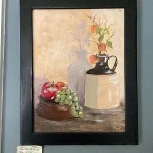 Framed still life painting featuring a cluster of green grapes, a red apple, a dark jug, and a cream