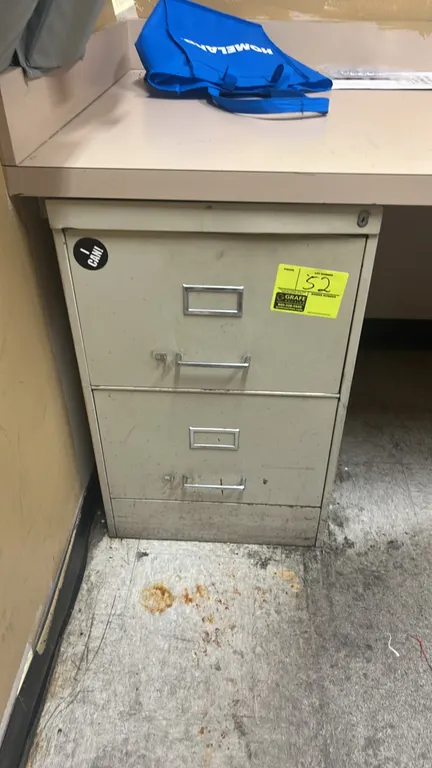 Metal 2-drawer vertical filing cabinet. Features pull handles, integrated label holders, and top lock cylinder. Desk and surrounding contents not incl