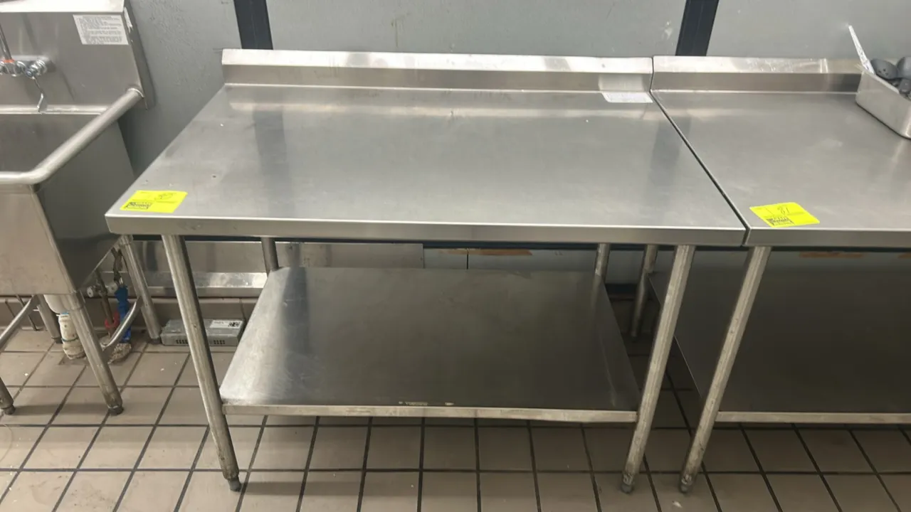 Stainless steel work table. Features a rear backsplash and lower undershelf. 48"W x 30"D. Mounted on tubular metal legs with adjustable bullet feet.