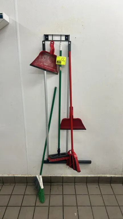 Assorted cleaning tools lot. Includes brooms, deck scrub brush, floor squeegee, upright lobby dustpan, hand dustpan, and wall-mounted storage rack.
