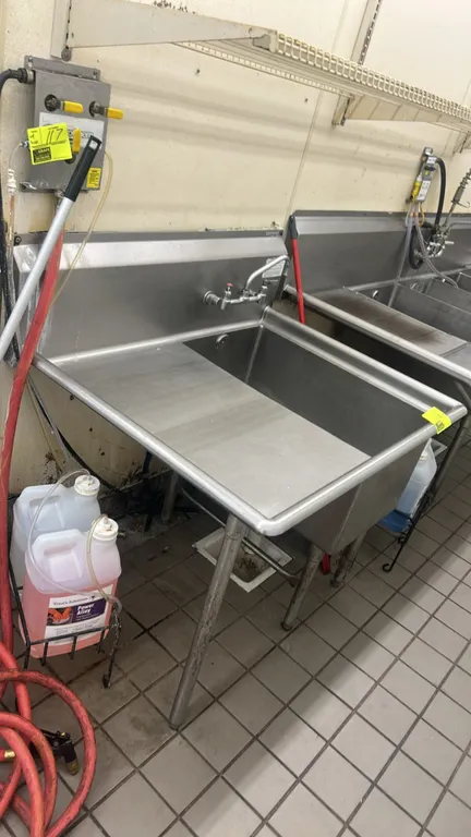 Stainless steel single-compartment sink. Features left-hand drainboard, backsplash, and splash-mount faucet. 44" x 36". Mounted on tubular legs with a