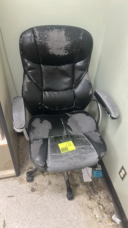 Cushioned office chair. Features black faux leather upholstery and padded armrests. Mounted on a 5-star base with casters. Note: Upholstery exhibits h