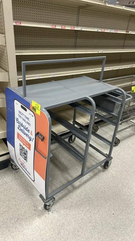 Metal mobile fulfillment cart. Features top shelf with overhead frame, slide rails for totes, side utility bin, end push handle, and side graphic pane