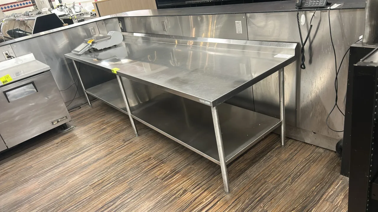 Stainless steel work table. Features a rear backsplash, full lower undershelf, and tubular legs with adjustable bullet feet. 10'L x 3'W. Scale and sur