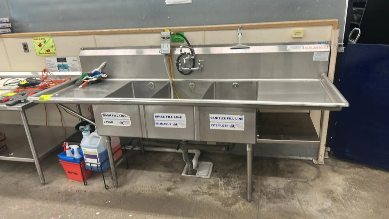 Stainless steel 3-compartment sink. Features left and right drainboards, integrated backsplash, splash-mount faucet, right lower undershelf, and a Hyd