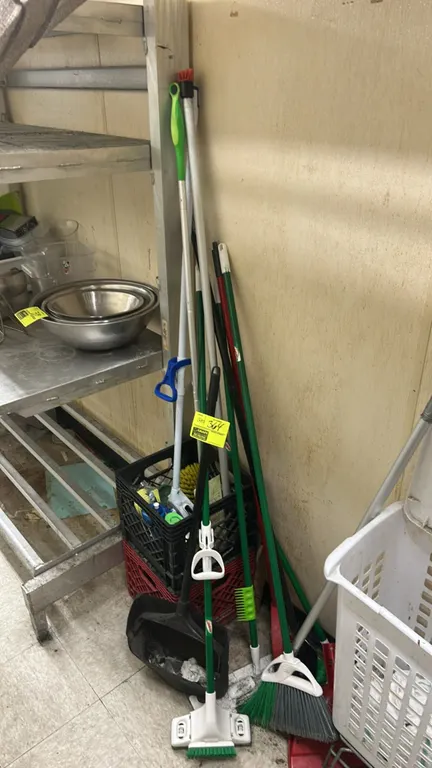 Assorted cleaning supplies and storage crates. Includes brooms, upright dust pans, scrub brushes, plastic milk crates, and remaining contents. Shelvin
