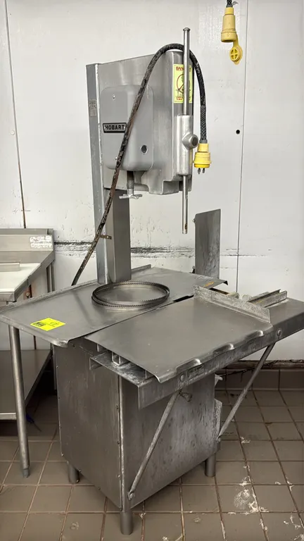 Hobart 5614 vertical meat bandsaw. Features stainless steel construction, sliding carriage table, stationary table, and removable scrap bin. Includes
