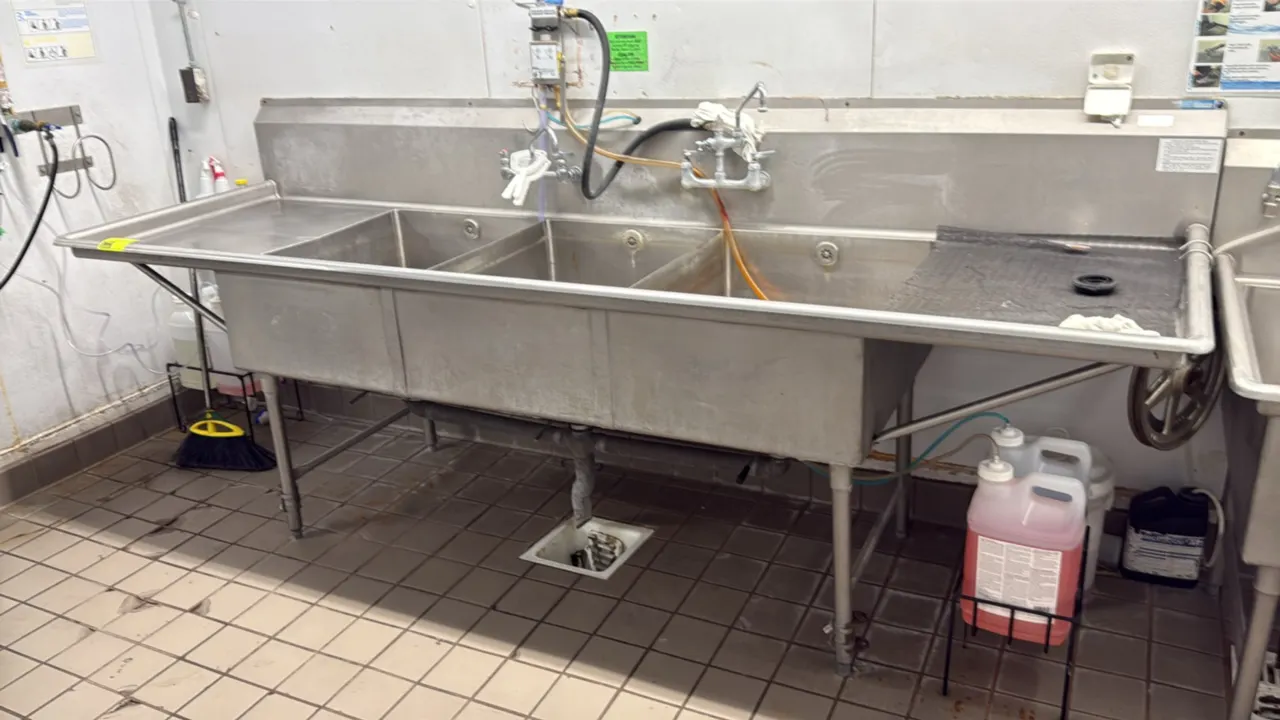 Stainless steel 3-compartment sink. Features left and right drainboards, high backsplash, and splash-mount faucet. 119" x 37". Mounted on tubular legs
