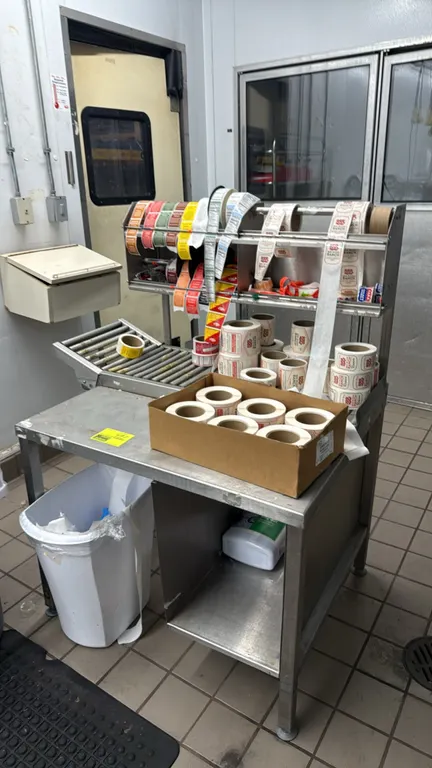 Stainless steel meat wrapping table. Features gravity roller conveyor track, 2-tier overhead label dispenser rack, lower open shelf, and assorted stic