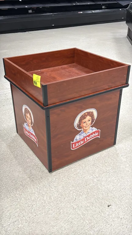Laminate merchandise dump display table. Features woodgrain laminate finish, raised perimeter bin sides, protective edge bumper trim, and Little Debbi