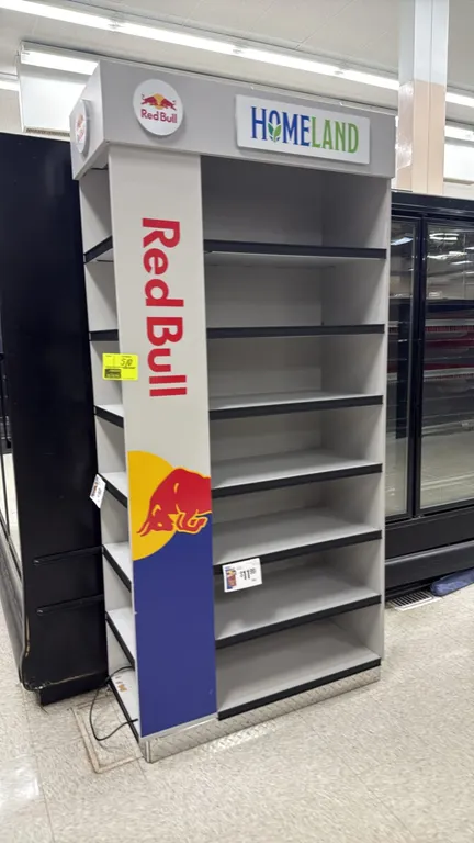 Red Bull merchandise display rack. Features Red Bull branding and graphics, Homeland header sign, 7 shelving levels, and diamond-plate base trim. 3'L.