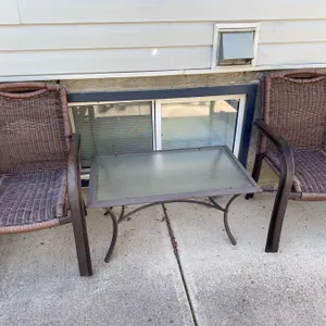 Outdoor patio set including a rectangular glass-top metal table and 4 brown wicker armchairs. Table 