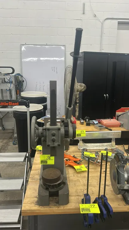 Manual ratchet-type arbor press. Features a ratcheting lever handle, side handwheel for rapid ram positioning, slotted anvil plate, and bench-mount ba