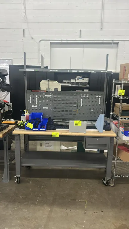 Portable metal workbench. Features wood top, lower storage shelf, single under-mount drawer, overhead shelf with dividers, and metal pegboard back pan