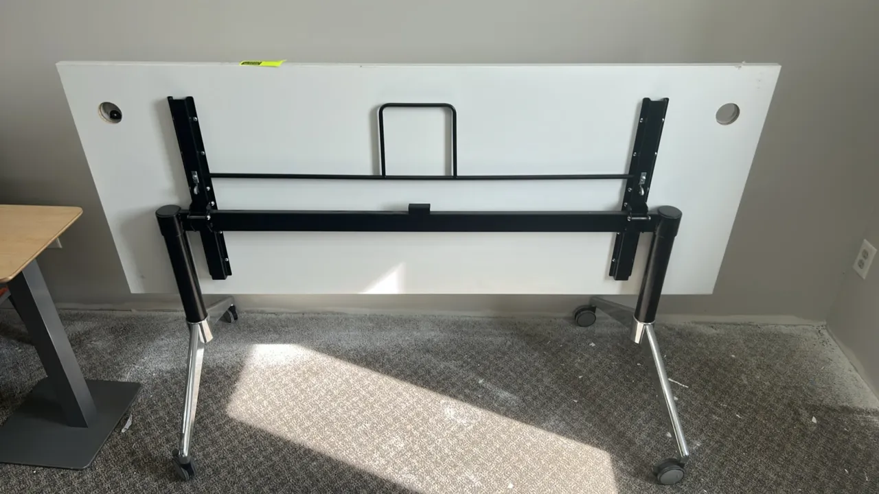 Collapsible flip-top table. Features a white laminate top with dual cable grommets and a metal folding frame. 71" x 28". Mounted on casters.