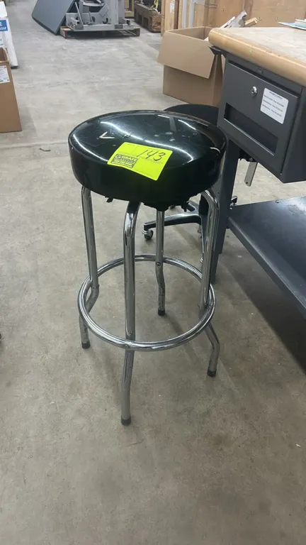 Cushioned shop stool. Features a black vinyl upholstered round seat and a tubular chrome metal frame with an integrated circular footrest ring. Note:
