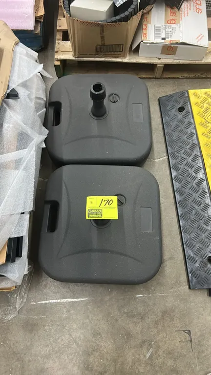 Fillable resin patio umbrella bases. Features black molded plastic construction, integrated carry handles, top fill caps, and central umbrella pole re