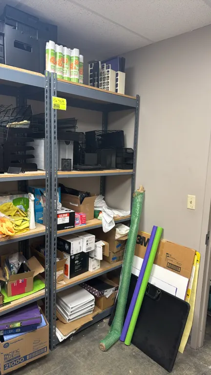 Metal 5-tier storage rack with contents. Features gray steel frame with composite decking. Includes assorted office and cleaning supplies such as desk