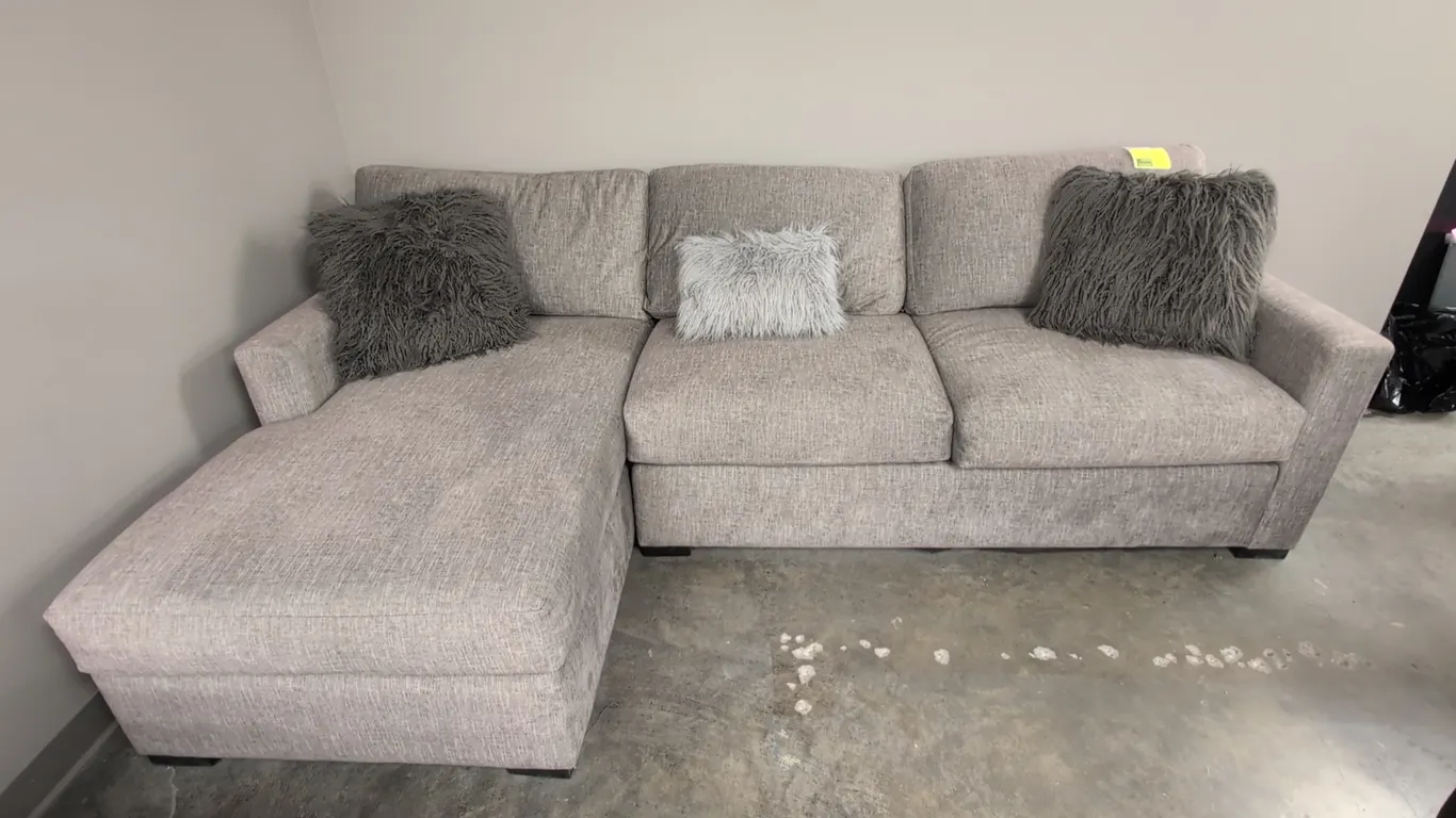 L-shaped sectional sofa. Features gray textured cloth upholstery, left-facing chaise lounge, cushioned seating, and (3) decorative shag throw pillows.