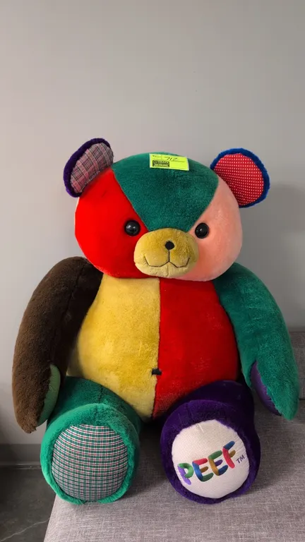 A large PEEF plush teddy bear. Features a multicolored patchwork plush design with patterned accents on the ears and foot, plastic safety eyes, and em