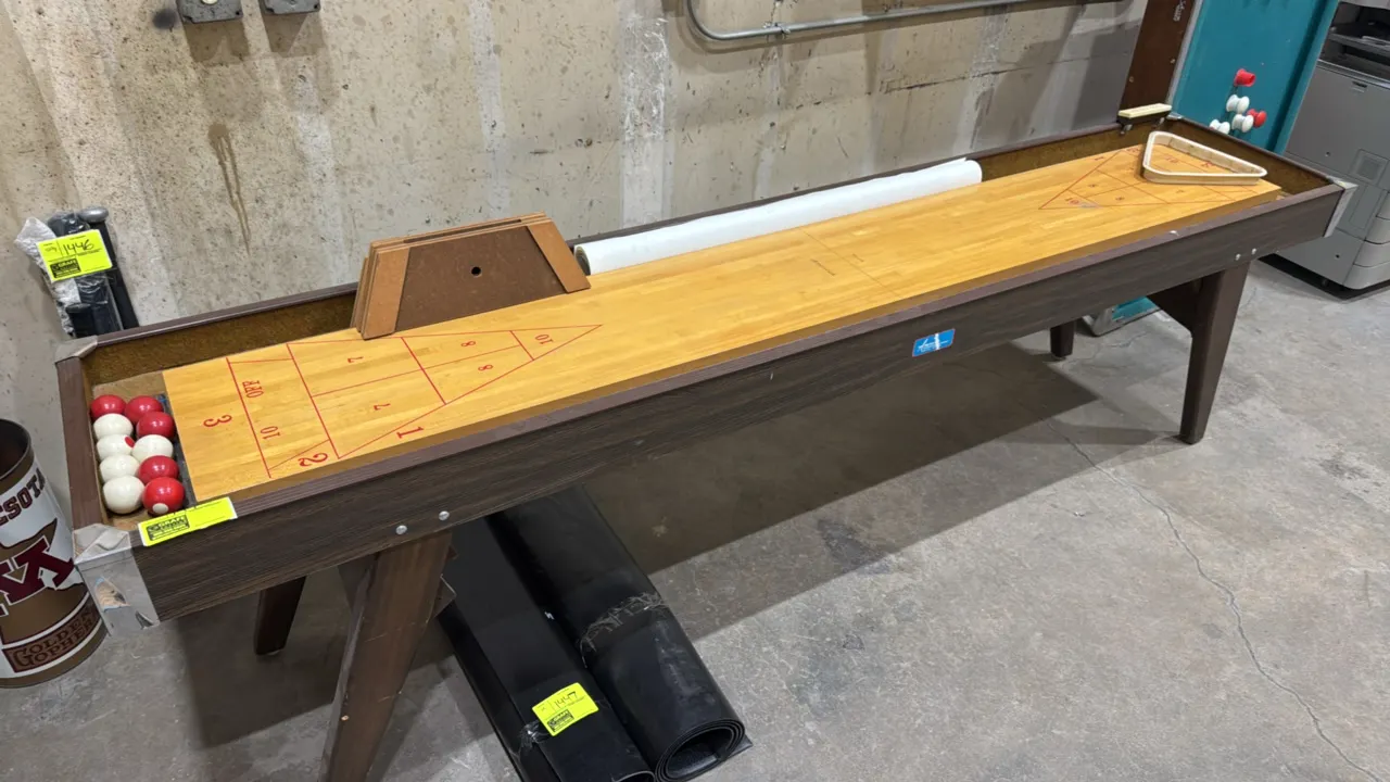 American Shuffleboard Company shuffleboard table. Features woodgrain laminate cabinet, wood playfield with printed red court markings, rail-mounted ma