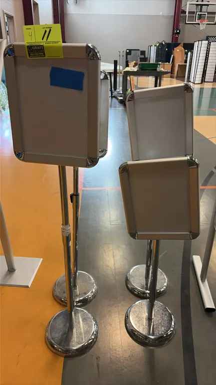 Adjustable sign stands. Features front-loading snap frames with rounded chrome corners and telescoping height-adjustable poles. Mounted on round chrom