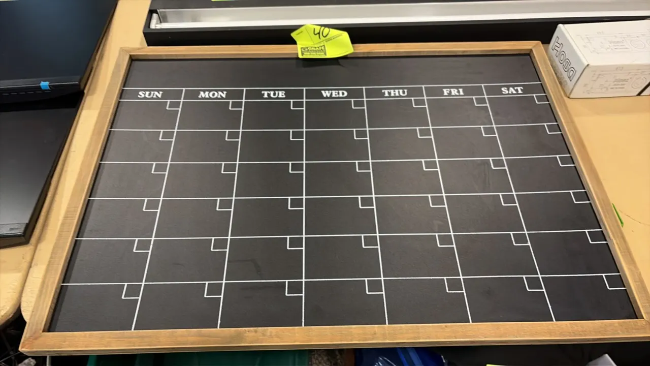 Framed chalkboard calendar. Features a monthly grid layout with printed days of the week (Sunday through Saturday), individual corner date blocks, and