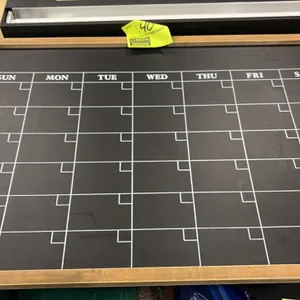 Framed chalkboard calendar. Features a monthly grid layout with printed days of the week (Sunday through Saturday), individual corner date blocks, and