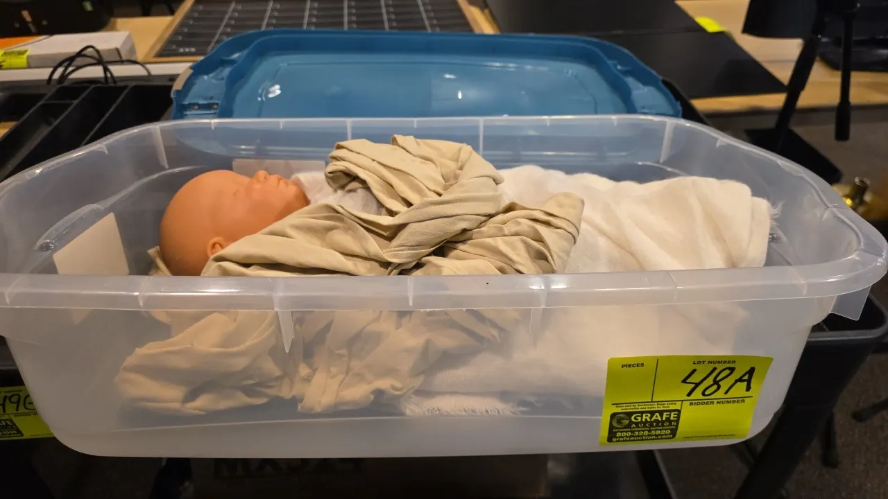 Plastic storage tote with baby doll. Includes fabric wrap and swaddling blanket. Clear poly storage container with blue lid.