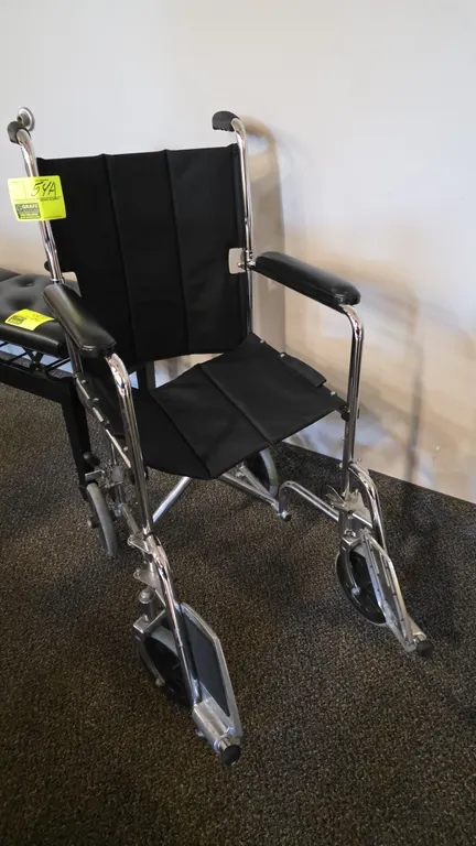 Transport wheelchair. Features a chrome-finished folding frame, black upholstery, padded armrests, fold-down backrest, and swing-away footrests. Equip
