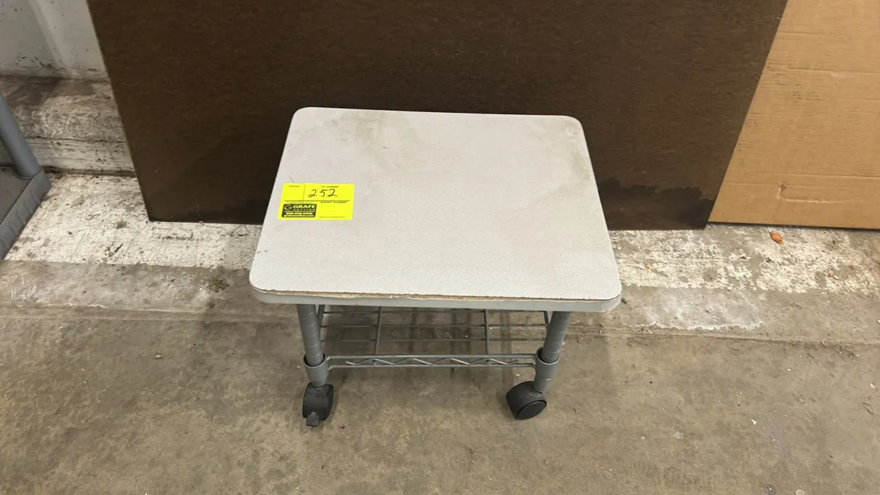 Rolling utility table. Features a gray laminate top, lower wire undershelf, and tubular metal frame. Mounted on swivel casters. Note: Front edge bandi