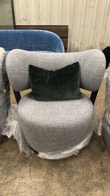 Adriana Hoyos Rumba upholstered lounge chair. Features gray textured fabric upholstery, curved backrest, black rear frame trim, and an included accent