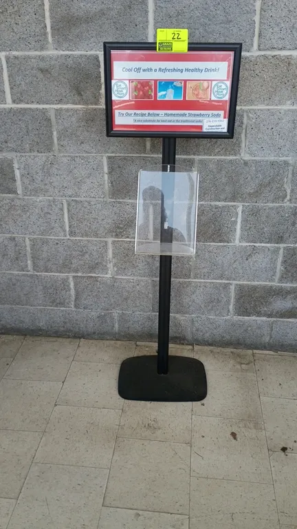 Metal sign holder. Features top sign frame, clear acrylic literature holder, and black metal post. 18" x 58". Mounted on a weighted base.