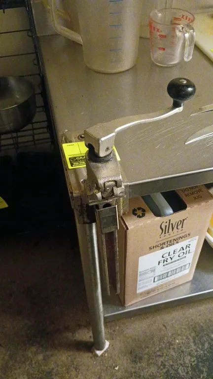 Commercial table-mount manual can opener. Features cast metal construction, hand crank with knob, and table-mount base plate. Table and surrounding it