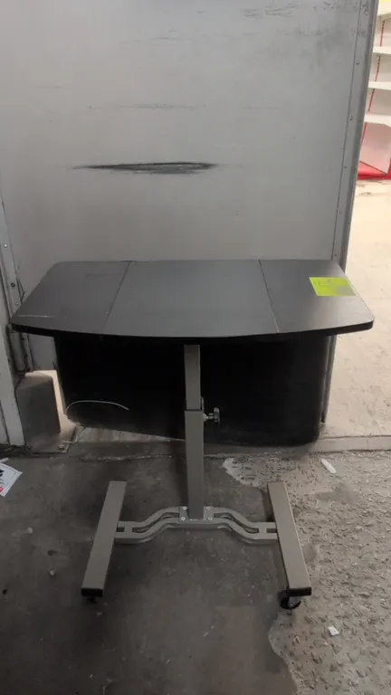 Portable laminate wood table. Features a height-adjustable column with tension adjustment knob and black laminate top. 26". Mounted on a painted metal