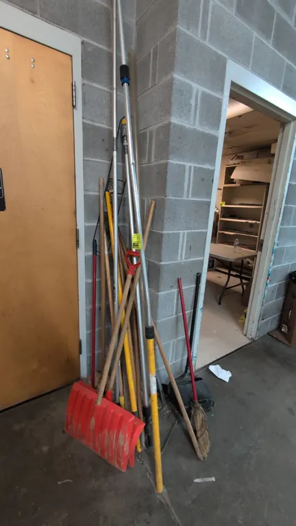 Assorted maintenance and cleaning tools. Includes plastic snow shovels, an upright lobby dustpan, broom, telescoping extension poles, and assorted woo