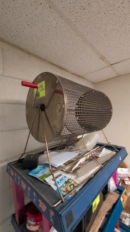 Metal raffle drum. Features perforated metal mesh drum, hinged ticket door, and manual hand crank with red grip handle. Mounted on a wire metal stand.