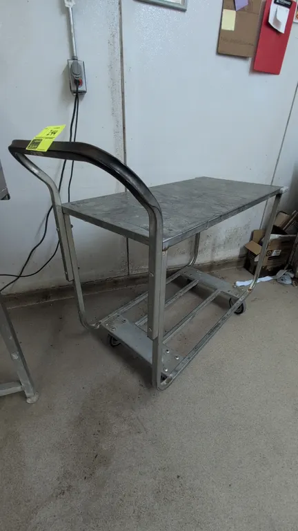 Metal platform stock cart. Features an upper solid metal deck, lower open-frame rack shelf, and tubular push handle. 44"L. Mounted on casters.