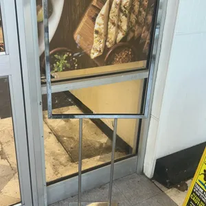 Metal floor-standing sign stand. Features a chrome-tone finish, top-loading poster frame, and dual upright supports. 2'W. Mounted on a flat rectangula