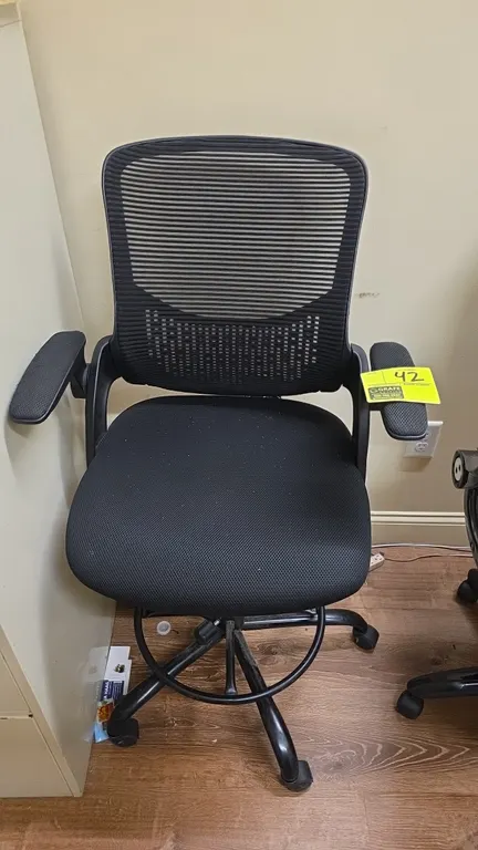 Mesh back drafting chair. Features black mesh back, fabric cushioned seat, flip-up armrests, and an adjustable circular footrest ring. Pneumatic heigh