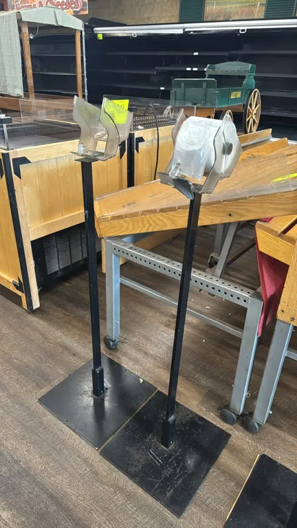 Metal produce bag stands. Features black metal uprights with square base plates and top-mounted clear plastic roll bag dispensers. Background fixtures