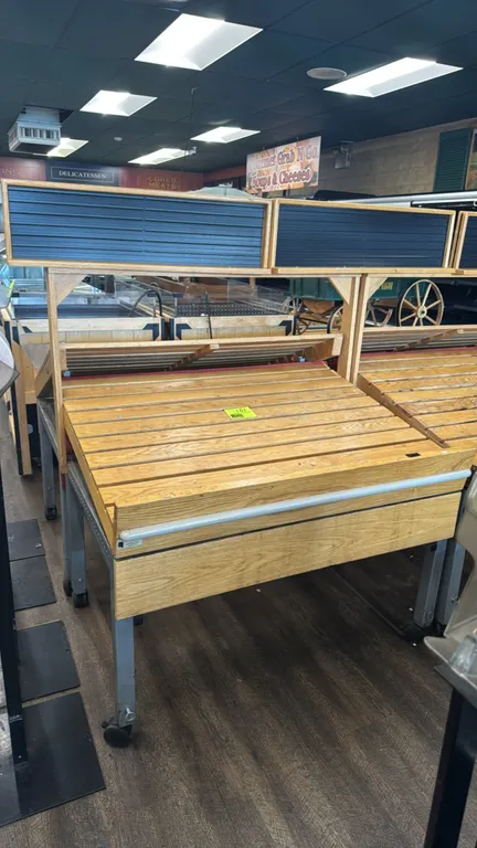 Tilting produce display tables. Features slatted wood display decks, adjustable tilt mechanisms, overhead display header boards, and front bumper rail