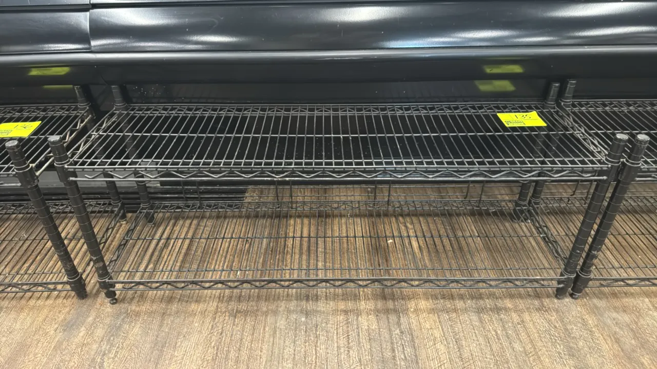 Black coated metal 2-tier wire rack. Features (2) wire shelves, lower shelf rear retaining ledge, and round tubular posts with adjustable leveling fee