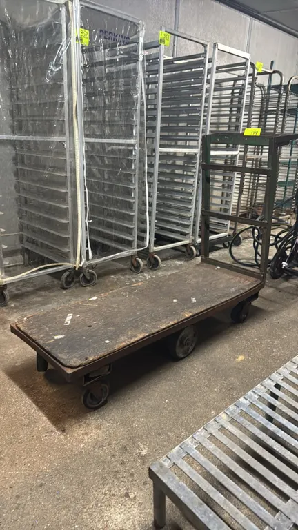 Metal 6-wheel platform cart with wood deck. Features an end push handle with upper shelf, steel frame, and wood platform. Mounted on 6 wheels with cen
