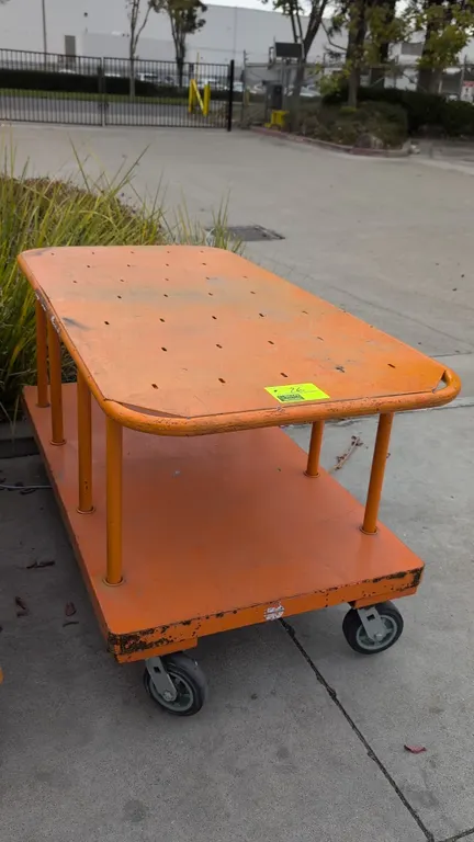 Two-tier metal platform cart. Features steel construction with orange finish and upper shelf with integrated tubular handles. Dimensions: 60"L x 30"W.