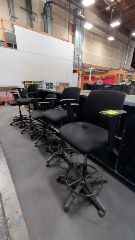 Adjustable drafting office chairs. Features black cloth upholstered cushioned seats and backrests, armrests, and circular foot rings. Mounted on 5-sta