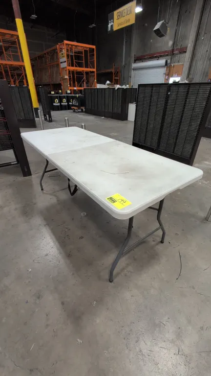 Poly fold-in-half folding table. Features a molded white plastic top and integrated carrying handle. Mounted on collapsible gray tubular metal legs. D
