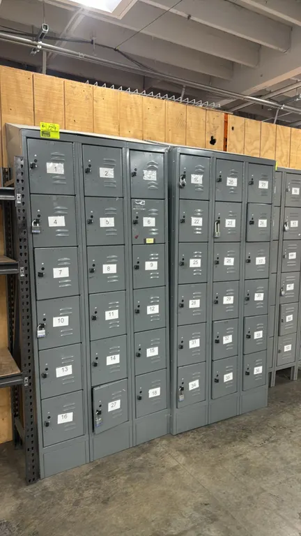 Metal locker sections. Features 6-tier box locker design per section, louvered steel doors with padlock hasps, label holders, and sloped tops. Gray fi